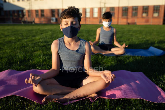 Zwei multiethnische Jungen mit Gesichtsmasken beim Yoga im Schulgarten. Grundschulbildung soziale Distanzierung der Gesundheitssicherheit während der Covid19 Coronavirus-Pandemie. — Stockfoto
