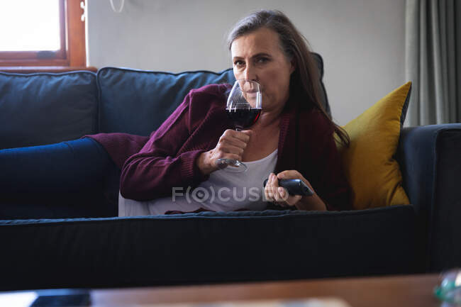 Mujer caucásica disfrutando del tiempo en casa, distanciamiento social y autoaislamiento en cuarentena, tumbada en el sofá en la sala de estar, bebiendo vino tinto, manteniendo el control remoto. - foto de stock