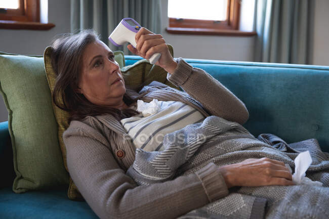 Хвора кавказька жінка проводить час удома, спілкуючись на відстані і самоізоляції в карантинній блокуванні, лежачи на дивані, вкритій ковдрою, тримаючи термометр, вимірюючи температуру.. — стокове фото