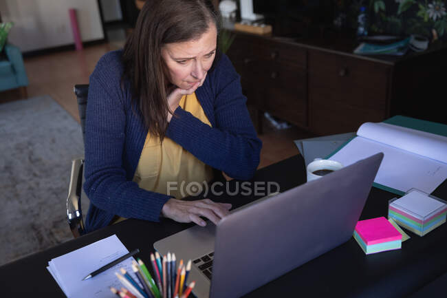 Mujer caucásica disfrutando del tiempo en casa, distanciamiento social y autoaislamiento en cuarentena, sentada en la mesa, usando una computadora portátil. - foto de stock