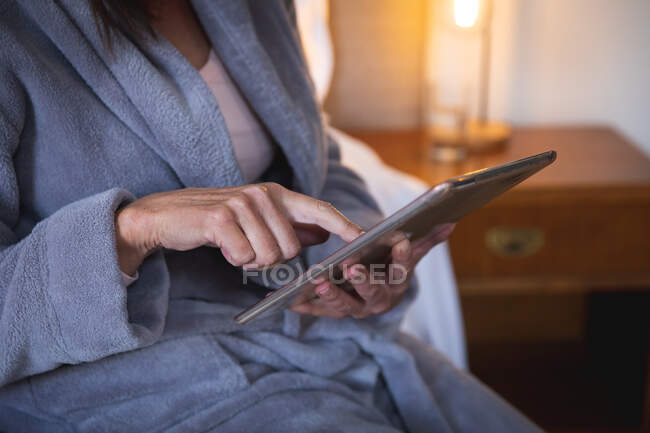 Section moyenne de la femme appréciant le temps à la maison, la distance sociale et l'isolement personnel en quarantaine verrouillée, assise sur le lit dans la chambre à coucher, à l'aide d'une tablette numérique. — Photo de stock