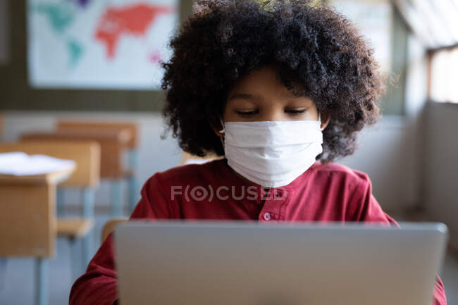 Хлопчик змішаної раси в масці для обличчя, використовуючи ноутбук, сидячи на своєму столі на уроці в школі. Початкова освіта соціальне дистанціювання охорона здоров'я під час пандемії коронавірусу Covid19 . — стокове фото