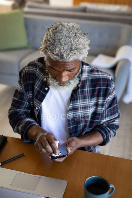 Un aîné afro-américain assis près d'une table, nettoyant un smartphone avec un mouchoir, prenant ses distances sociales et s'isolant en quarantaine — Photo de stock