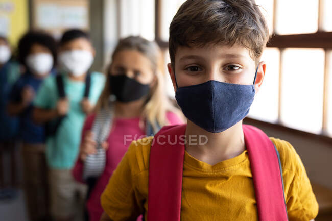 Groupe multi-ethnique d'élèves du primaire regardant la caméra, portant des masques dans le hall de l'école. Enseignement primaire distanciation sociale sécurité sanitaire pendant la pandémie de coronavirus Covid19. — Photo de stock