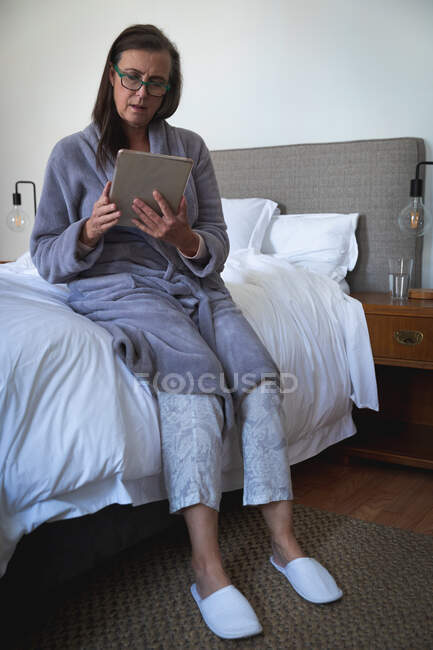 Femme caucasienne appréciant le temps à la maison, la distance sociale et l'isolement personnel en quarantaine verrouillée, assise sur le lit dans la chambre, à l'aide d'une tablette numérique. — Photo de stock