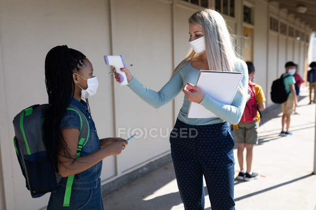 Кавказька вчителька в масці обличчя вимірює температуру дівчинки в початковій школі. Первинна освіта Соціальна безпека для здоров'я під час пандемії Ковіда19 Коронавірус. — стокове фото