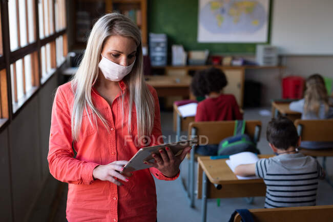 Maestra caucásica usando mascarilla usando tableta digital en la escuela. Educación primaria distanciamiento social seguridad sanitaria durante la pandemia del Coronavirus Covid19. - foto de stock