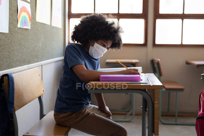 Mixte garçon de race assis au bureau portant un masque dans la salle de classe. Enseignement primaire distanciation sociale sécurité sanitaire pendant la pandémie de coronavirus Covid19. — Photo de stock