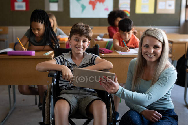 Porträt eines behinderten kaukasischen Jungen, der im Rollstuhl sitzt, und seiner Lehrerin mit Tablet im Klassenzimmer. Grundschulbildung soziale Distanzierung der Gesundheitssicherheit während der Covid19 Coronavirus-Pandemie. — Stockfoto