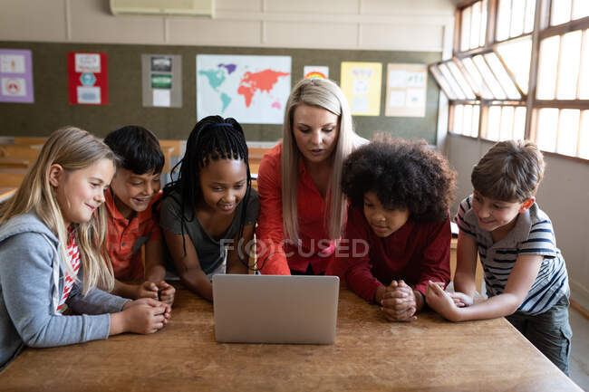 Insegnante caucasica femminile e gruppo multietnico di bambini che utilizzano il computer portatile durante la lezione. Istruzione primaria distanza sociale sicurezza sanitaria durante la pandemia di Covid19 Coronavirus. — Foto stock