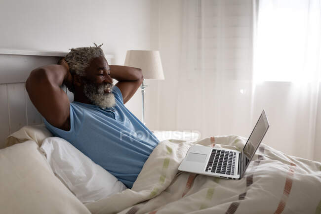 Старший афроамериканец лежит на кровати в спальне, используя ноутбук и улыбаясь, социальное дистанцирование и самоизоляция в карантинной изоляции — стоковое фото