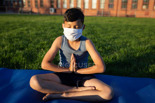 Mischlingsjunge mit Gesichtsmaske beim Yoga im Schulgarten. Grundschulbildung soziale Distanzierung der Gesundheitssicherheit während der Covid19 Coronavirus-Pandemie. — Stockfoto
