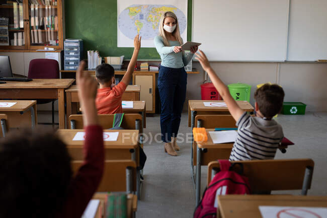 Група з декількох етнічних дітей, які сидять на своєму столі під час уроку з вчителем-жінкою в масці для обличчя. Початкова освіта соціальне дистанціювання охорона здоров'я під час пандемії коронавірусу Covid19 . — стокове фото