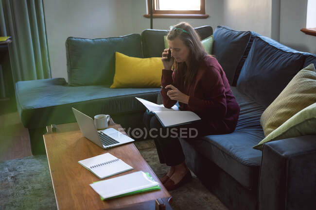 Кавказька жінка, яка проводить час удома, спілкується на відстані і самотності в карантині, сидить на дивані в вітальні, користується ноутбуком, розмовляє по телефону.. — стокове фото