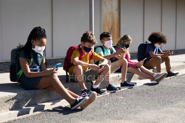 Groupe multi ethnique d'enfants de l'école primaire portant des masques faciaux à l'aide de smartphones assis ensemble. Enseignement primaire distanciation sociale sécurité sanitaire pendant la pandémie de coronavirus Covid19 — Photo de stock