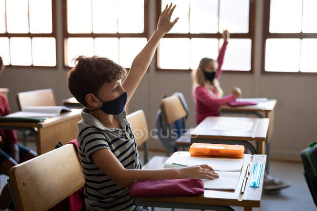 Grupo de niños multiétnicos que usan máscaras faciales, sentados en su escritorio durante la lección. Educación primaria distanciamiento social seguridad sanitaria durante la pandemia del Coronavirus Covid19. - foto de stock