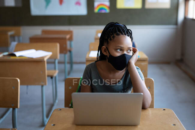Chica de raza mixta con una máscara facial, el uso de un ordenador portátil mientras está sentado en su escritorio en la clase en la escuela. Educación primaria distanciamiento social seguridad sanitaria durante la pandemia del Coronavirus Covid19. - foto de stock