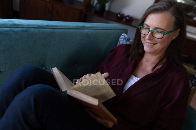 Kaukasische Frau genießt Zeit zu Hause, soziale Distanzierung und Selbstisolierung in Quarantäne, liegt auf dem Sofa im Wohnzimmer, liest ein Buch. — Stockfoto