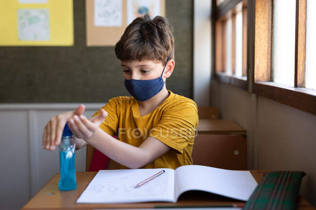 Kaukasischer Junge mit Mundschutz, der seine Hände desinfiziert, während er auf ihrem Schreibtisch im Klassenzimmer sitzt. Grundschulbildung soziale Distanzierung der Gesundheitssicherheit während der Covid19 Coronavirus-Pandemie. — Stockfoto