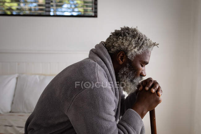 Hombre mayor afroamericano sentado en una cama en un dormitorio, descansando su cabeza sobre un bastón, distanciamiento social y aislamiento en cuarentena - foto de stock