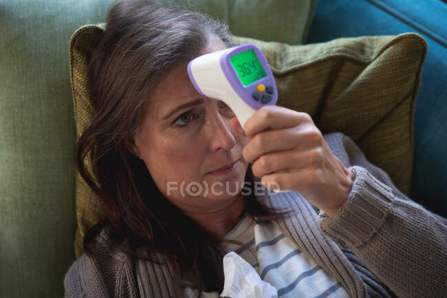 Больная белая женщина проводит время дома, социальное дистанцирование и самоизоляция в карантинной изоляции, лежит на диване, измеряет температуру, держит термометр. — стоковое фото