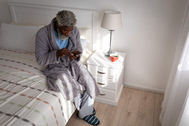 Homme âgé afro-américain assis sur un lit dans une chambre à coucher, en utilisant un smartphone, la distance sociale et l'isolement personnel en quarantaine verrouillage — Photo de stock