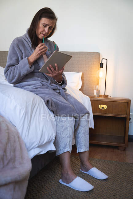 Кавказька жінка, яка проводить час удома, спілкується на відстані і самотності в карантині, сидить на ліжку в спальні, користуючись цифровою табличкою.. — стокове фото