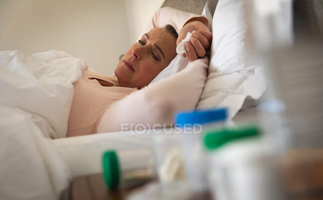 Mujer caucásica enferma pasando tiempo en casa, distanciamiento social y aislamiento en cuarentena, acostada en la cama, sosteniendo tejido. - foto de stock