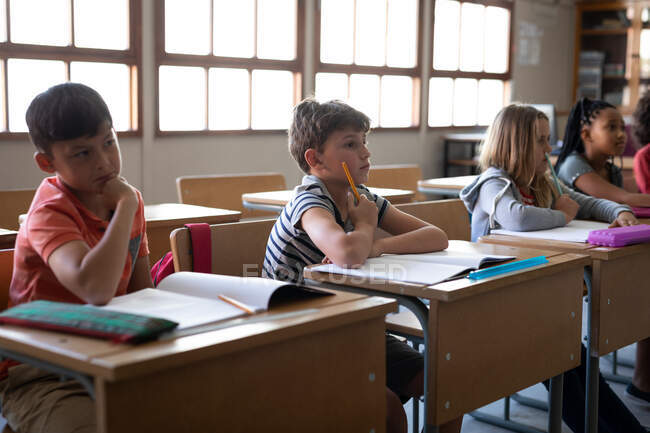Groupe d'enfants multiethniques assis sur leur bureau dans la salle de classe à l'école. Enseignement primaire distanciation sociale sécurité sanitaire pendant la pandémie de coronavirus Covid19. — Photo de stock