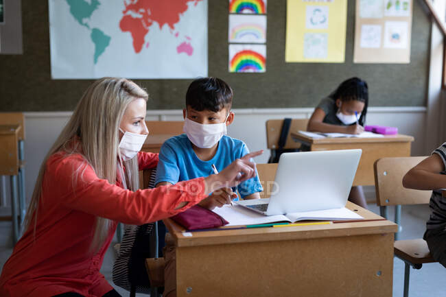 Female Caucasian teacher and mixed race boy wearing face masks using laptop in class at school. Primary education social distancing health safety during Covid19 Coronavirus pandemic. — Stock Photo