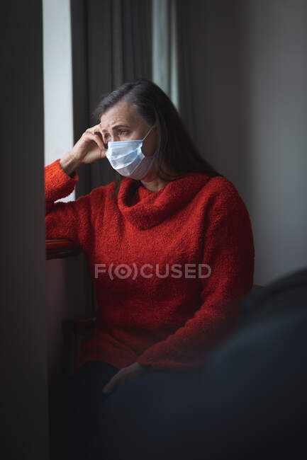 Mujer caucásica preocupada pasando tiempo en casa, distanciamiento social y aislamiento en cuarentena, usando mascarilla facial para protegerse de la infección por el coronavirus Covid 19, mirando por la ventana. - foto de stock