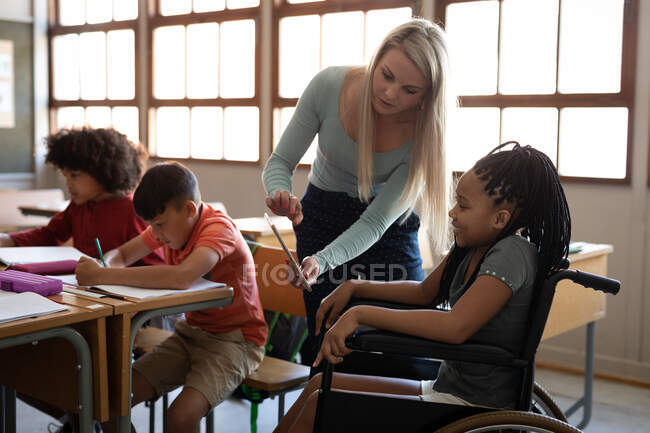 Deshabilitar chica de raza mixta sentado en su silla de ruedas y su maestra usando tableta en el aula. Educación primaria distanciamiento social seguridad sanitaria durante la pandemia del Coronavirus Covid19. - foto de stock