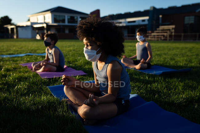 Gruppo di bambini multietnici che indossano maschere facciali che praticano yoga nel giardino della scuola. Istruzione primaria distanza sociale sicurezza sanitaria durante la pandemia di Covid19 Coronavirus. — Foto stock