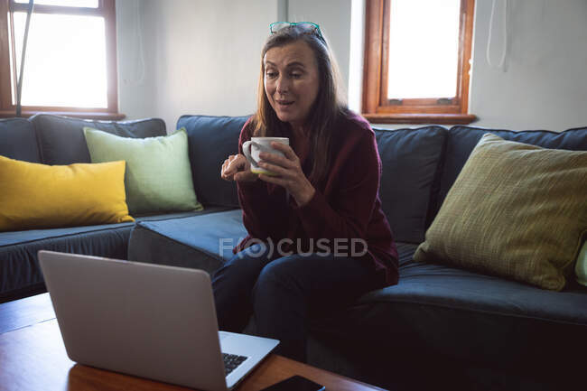 Mujer caucásica disfrutando del tiempo en casa, distanciamiento social y autoaislamiento en cuarentena, sentada en un sofá en la sala de estar, usando una computadora portátil, teniendo videollamada. - foto de stock