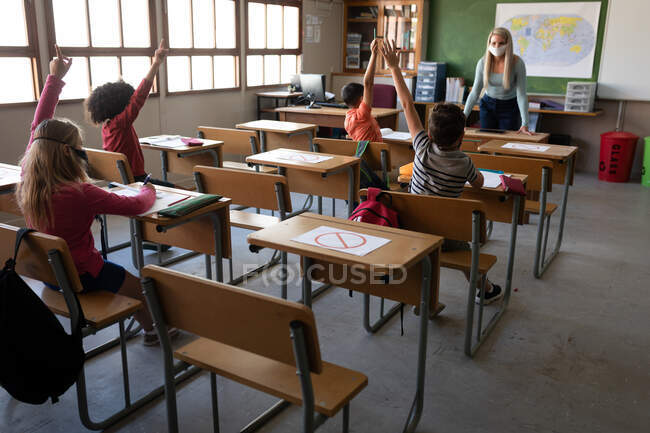 Groupe d'enfants multiethniques assis sur leur bureau pendant la leçon avec une enseignante portant un masque facial. Enseignement primaire distanciation sociale sécurité sanitaire pendant la pandémie de coronavirus Covid19. — Photo de stock