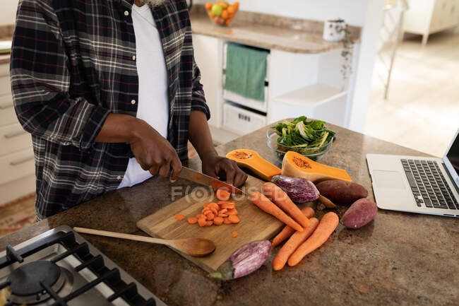 Hombre de pie en una cocina, cortando verduras con un cuchillo, distanciamiento social y autoaislamiento en cuarentena - foto de stock