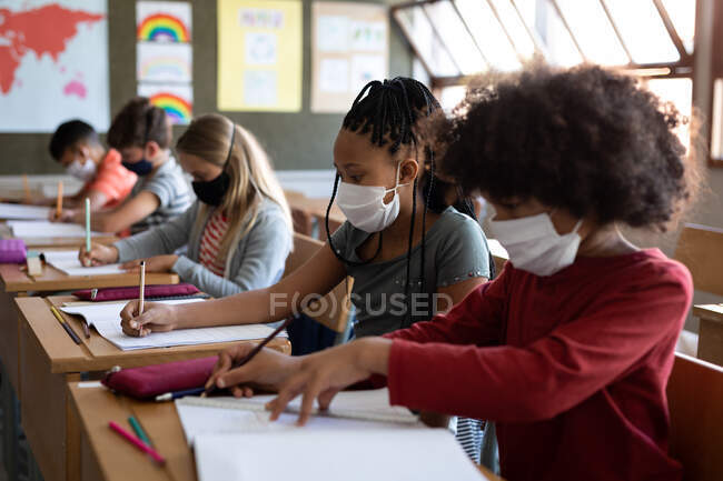 Groupe d'enfants multi-ethniques portant des masques faciaux tout en étudiant en classe à l'école. Enseignement primaire distanciation sociale sécurité sanitaire pendant la pandémie de coronavirus Covid19. — Photo de stock