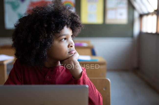 Nachdenklicher Mischlingsjunge mit Laptop, während er in der Schule auf seinem Schreibtisch sitzt. Grundschulbildung soziale Distanzierung der Gesundheitssicherheit während der Covid19 Coronavirus-Pandemie. — Stockfoto