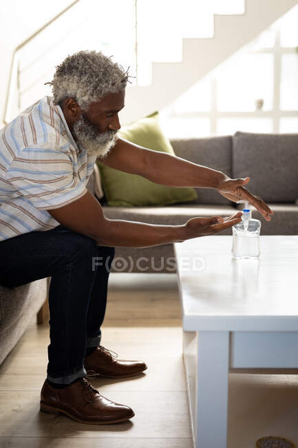 Hombre mayor afroamericano sentado en un sofá, vertiendo jabón en sus manos, distanciamiento social y aislamiento en cuarentena - foto de stock