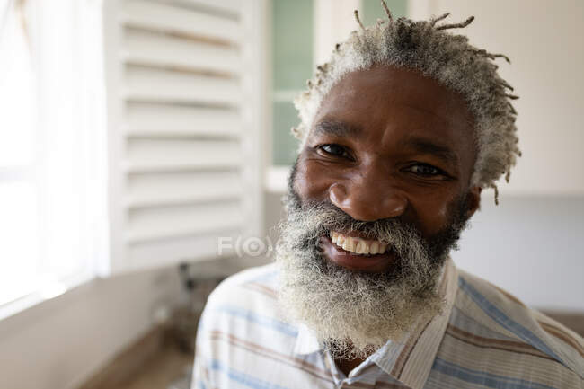 Retrato de um homem idoso afro-americano em pé em um quarto, olhando para a câmera e sorrindo, distanciamento social e auto-isolamento em quarentena — Fotografia de Stock