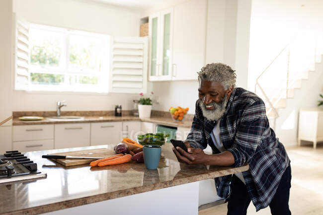 Homme âgé afro-américain debout dans une cuisine, en utilisant un smartphone, la distance sociale et l'isolement personnel en quarantaine verrouillage — Photo de stock