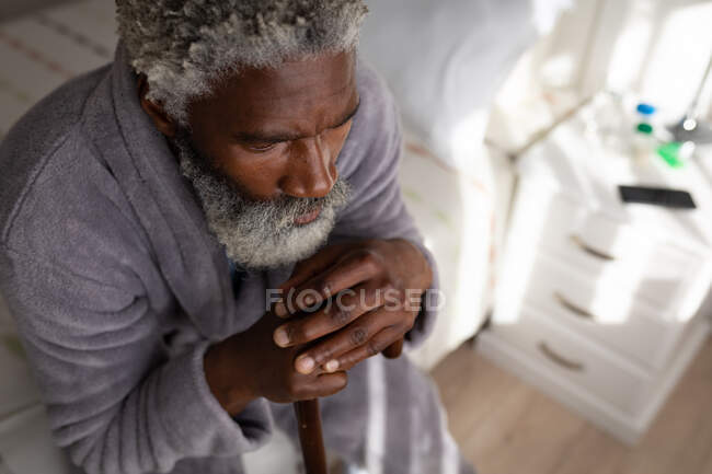 Afroamerikanischer Senior sitzt auf einem Bett im Schlafzimmer, ruht seinen Kopf auf einem Stock, soziale Distanzierung und Selbstisolierung in Quarantäne — Stockfoto