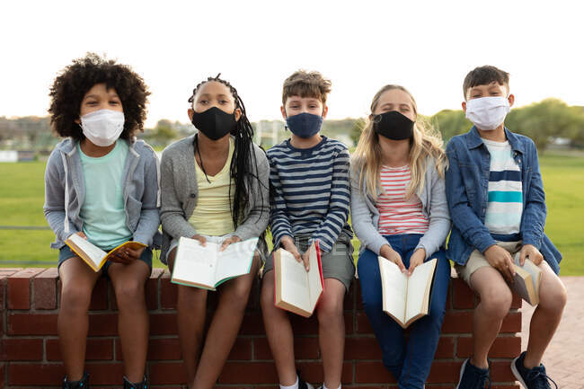 Groupe d'enfants multiethniques portant des masques faciaux lisant des livres assis sur le mur pendant une pause. Enseignement primaire distanciation sociale sécurité sanitaire pendant la pandémie de coronavirus Covid19. — Photo de stock