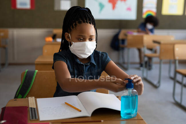 Menina de raça mista usando máscara facial, higienizando as mãos enquanto estava sentada em sua mesa na sala de aula. Educação primária distanciamento social segurança sanitária durante Covid19 pandemia de coronavírus. — Fotografia de Stock
