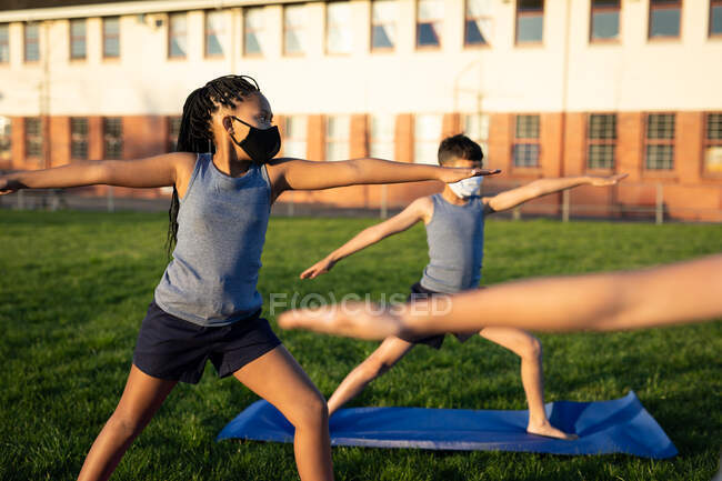 Group of multi ethnic kids wearing face masks performing yoga in the school garden. Primary education social distancing health safety during Covid19 Coronavirus pandemic. — Stock Photo