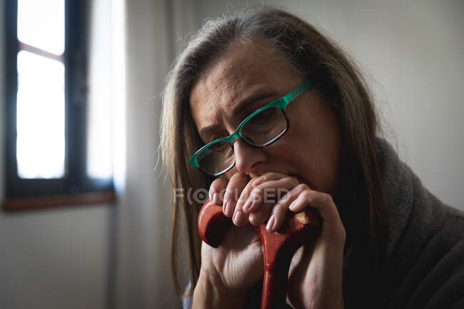 Mujer caucásica preocupada pasando tiempo en casa, distanciamiento social y autoaislamiento en cuarentena, usando gafas, sosteniendo y apoyándose en bastón. - foto de stock