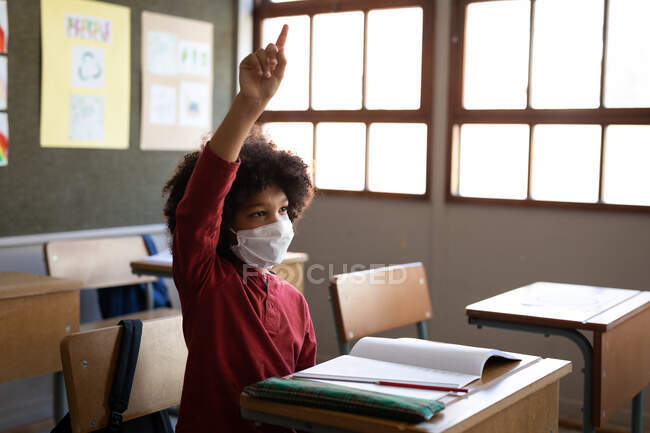 Niño de raza mixta con máscara facial, sentado en su escritorio durante la lección. Educación primaria distanciamiento social seguridad sanitaria durante la pandemia del Coronavirus Covid19. - foto de stock