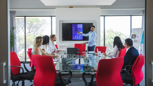 Homme d'affaires mixte debout à côté d'un écran donnant une présentation à des collègues masculins et féminins assis dans une salle de réunion. Professionnels créatifs d'affaires travaillant dans un bureau moderne occupé. — Photo de stock