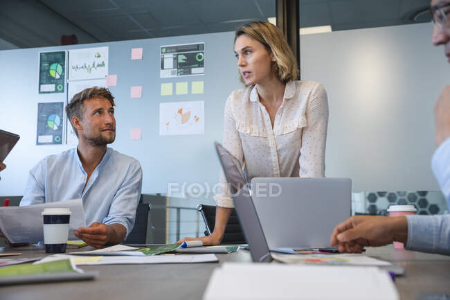 Caucasian businesswoman standing to address colleagues during a team meeting, a Caucasian male colleague sitting beside her listening. Creative business professionals working in a busy modern office. — Stock Photo