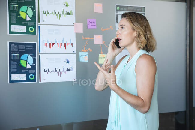 Mulher de negócios caucasiana casualmente vestida inteligente falando no smartphone, um conselho com gráficos e informações sobre isso em segundo plano. Profissional de negócios criativos trabalhando em um escritório moderno ocupado. — Fotografia de Stock